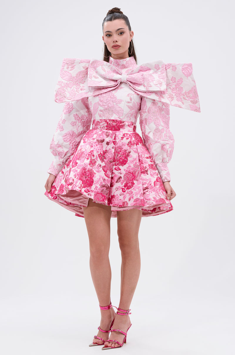 A woman wears the RENAISSANCE TWO-TONE BROCADE MINI DRESS IN PINK, featuring voluminous long sleeves, a sculptural chest bow, and a flared skirt. Her hair is tied up and she pairs the dress with pink high-heeled sandals.