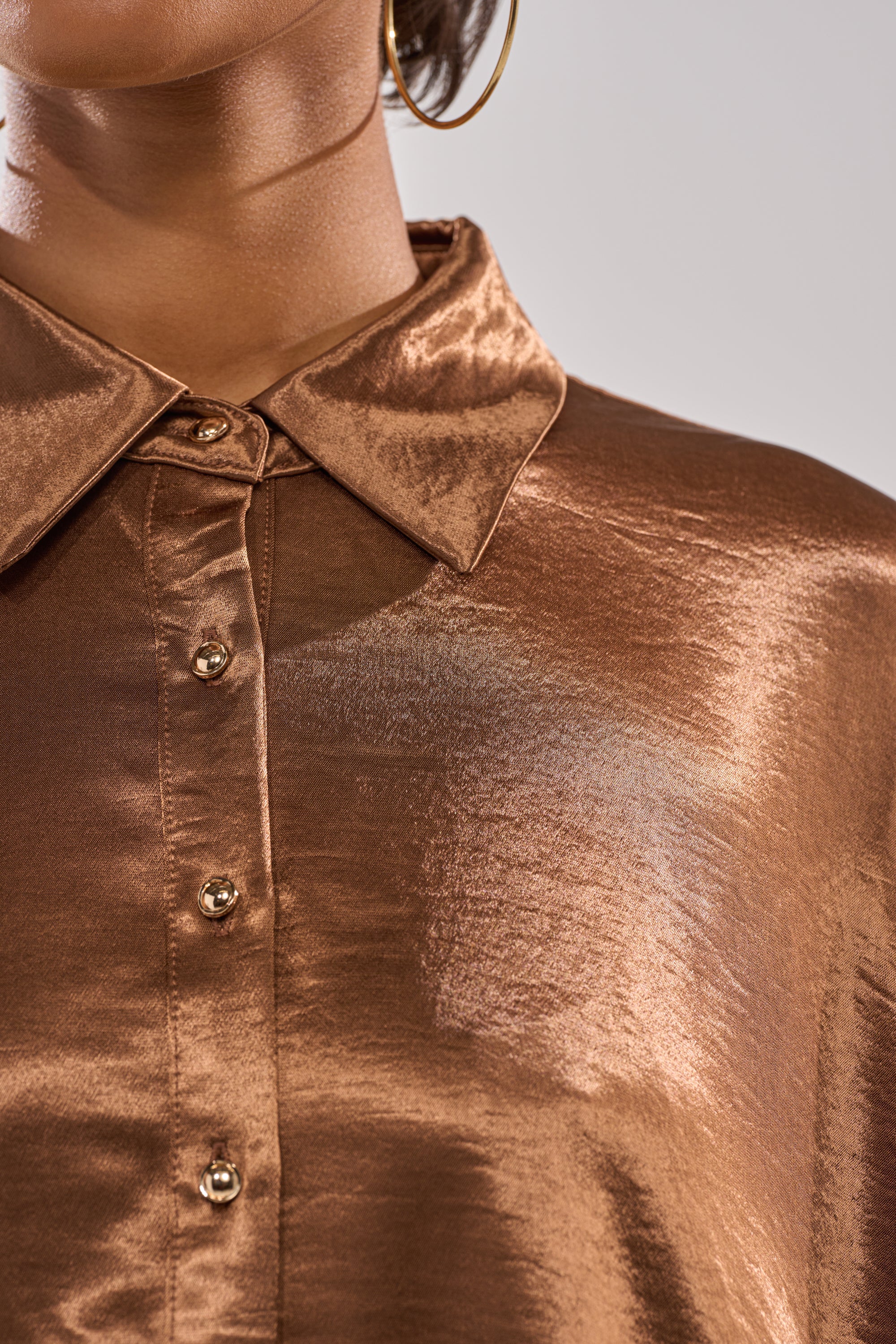 Close-up of a medium brown-skinned person wearing the WONDERLUST SATIN LONG SLEEVE MINI DRESS IN BROWN, featuring gold buttons and a pointed collar, styled with a large gold hoop earring for a timeless, elegant look. 8