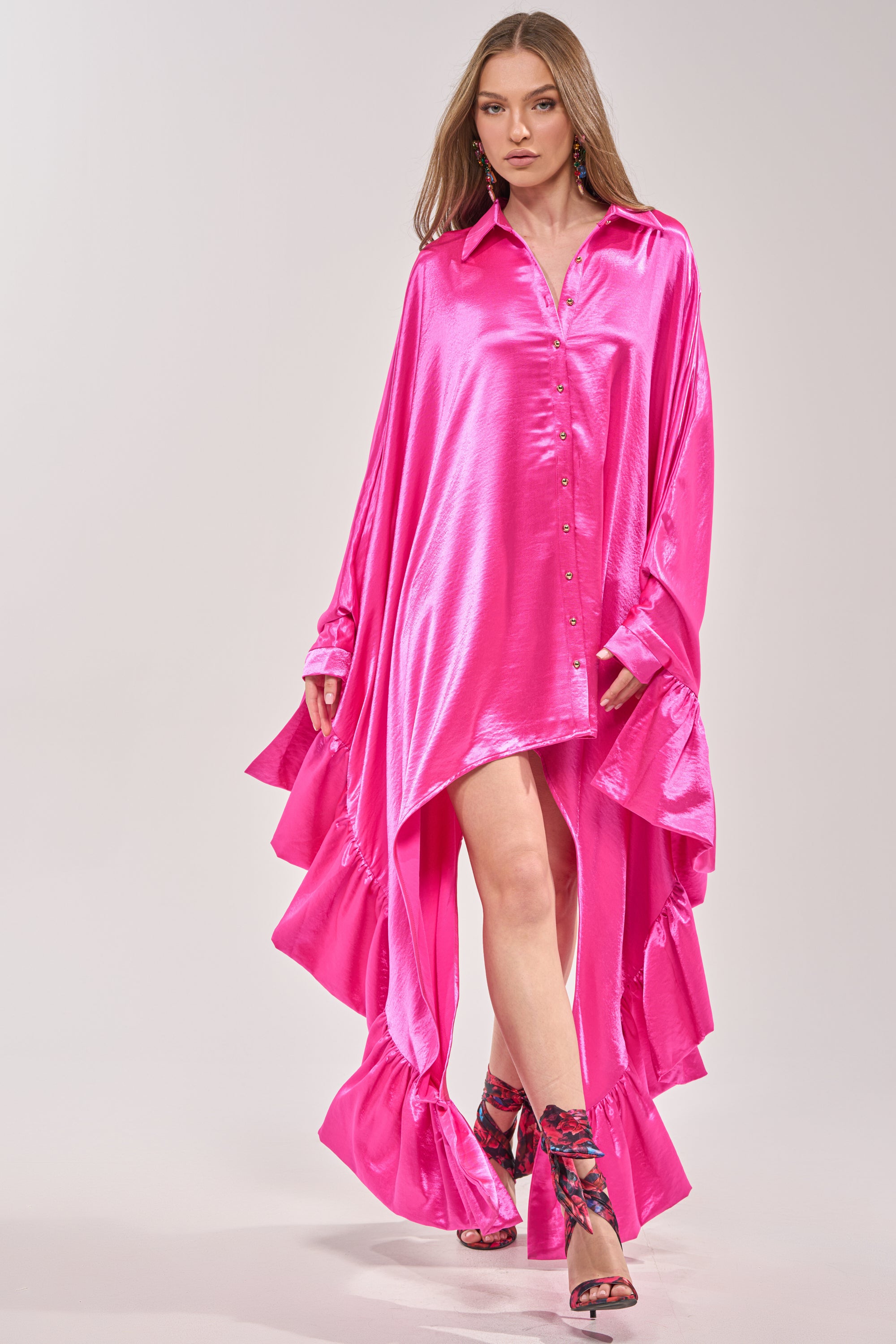 A woman walks confidently in the WONDERLUST SATIN LONG SLEEVE MINI DRESS IN PINK, featuring a ruffled asymmetrical hem, paired with floral high-heeled sandals against a light background. 3