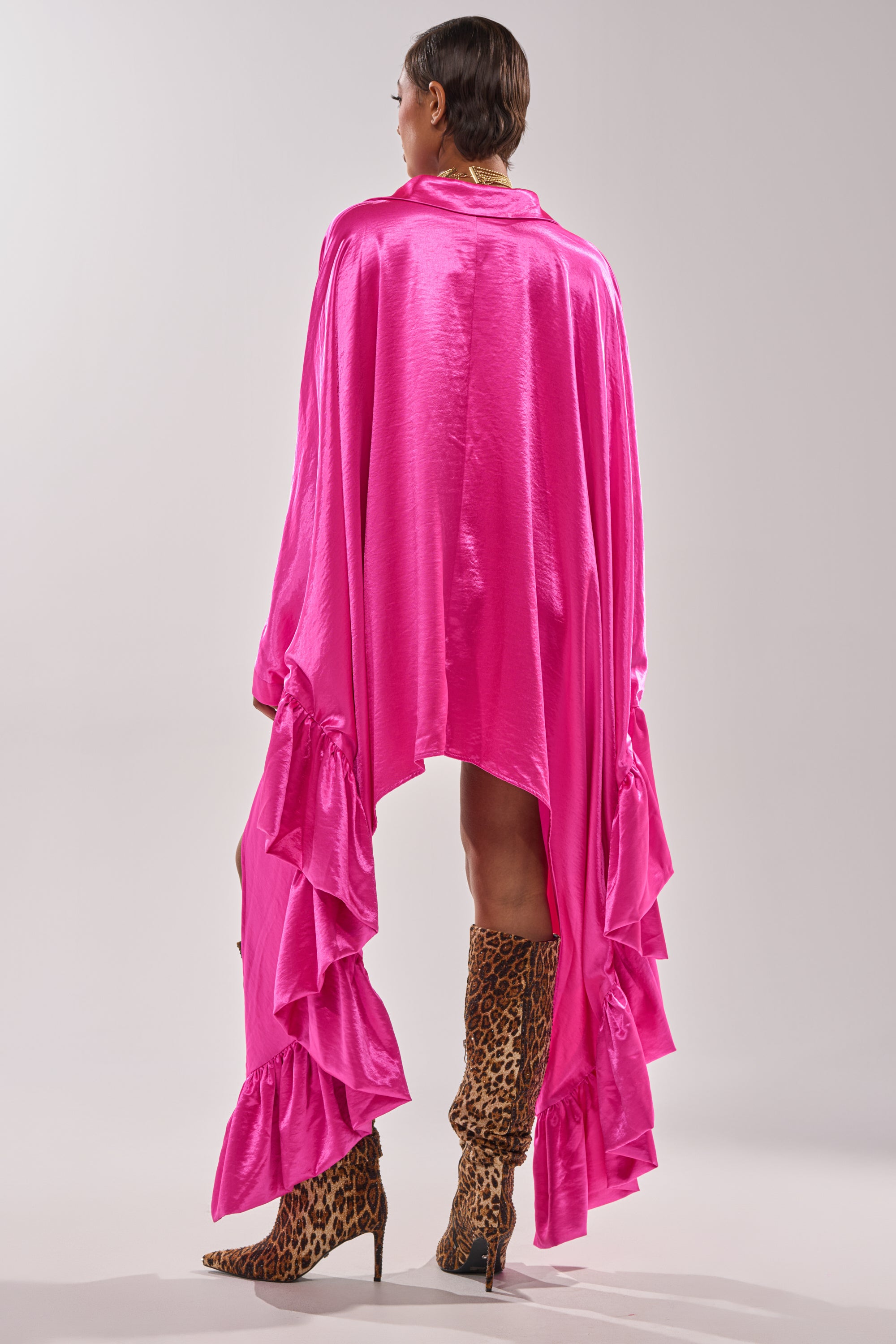 A person faces away, wearing the WONDERLUST Satin Long Sleeve Mini Dress in Pink with ruffled details and shiny finish, styled with knee-high leopard print heeled boots and short dark hair. 5