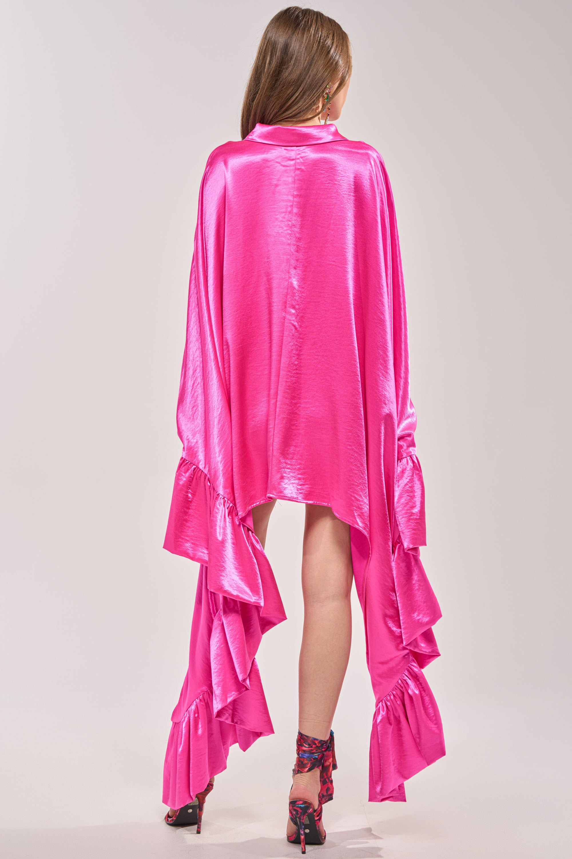 A person faces away, wearing the WONDERLUST Satin Long Sleeve Mini Dress in bright pink with striking ruffled cuffs and hem. Their straight hair is down, paired with floral high heels against a plain light backdrop. 5