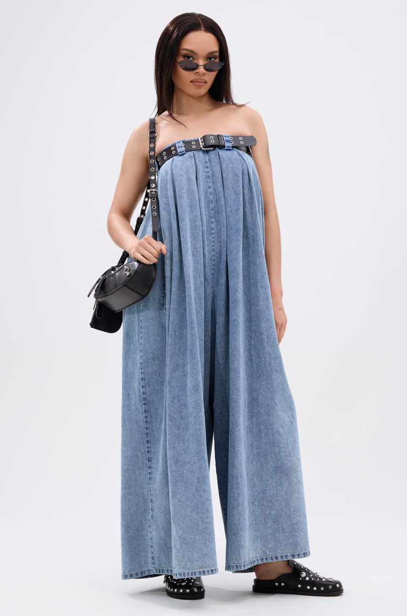 A woman poses in the DIANE STRAPLESS OVERSIZED JUMPSUIT IN DENIM, styled with a faux leather belt, black studded sandals, a black shoulder bag, and small sunglasses against a plain white background.