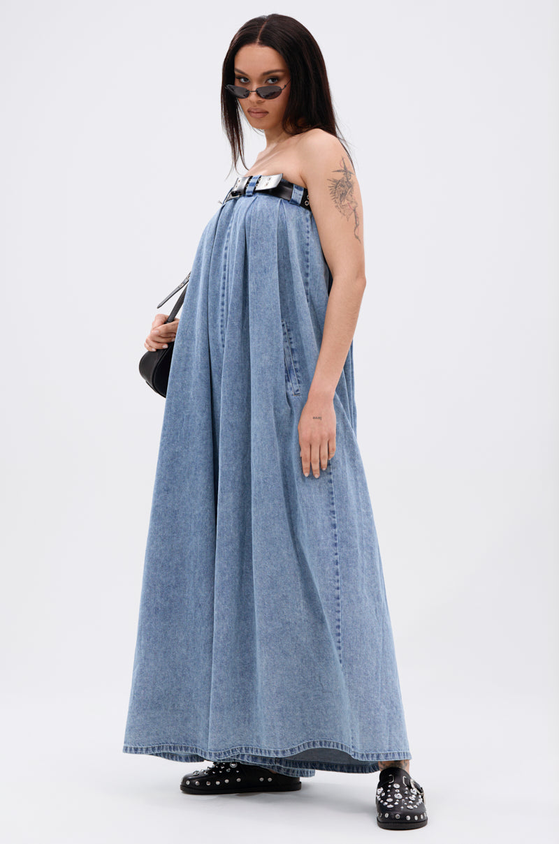 A person wears the DIANE STRAPLESS OVERSIZED JUMPSUIT IN DENIM with a faux leather belt, round sunglasses, black studded shoes, and a black bag against a plain white background. They have an upper arm tattoo.
