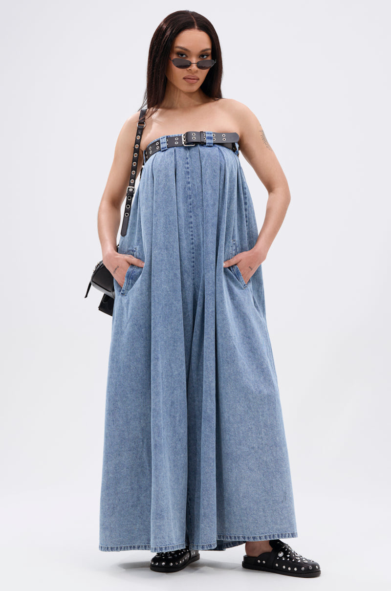 A woman poses with her hands in the pockets, wearing the DIANE STRAPLESS OVERSIZED JUMPSUIT IN DENIM paired with a faux leather belt, black studded shoes, a black shoulder bag, and sunglasses against a plain white background.