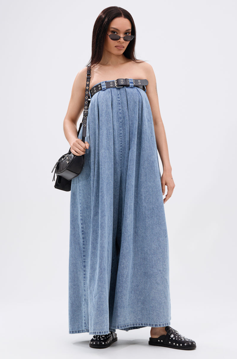 A woman in the DIANE STRAPLESS OVERSIZED JUMPSUIT IN DENIM, styled with a black belt, black handbag, round sunglasses, and black studded slip-on shoes, stands against a plain white background.