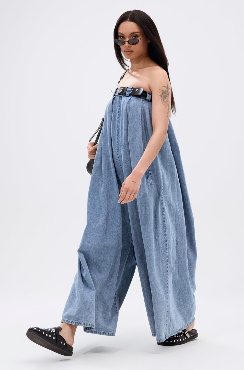 A woman confidently showcases the effortless style of the DIANE STRAPLESS OVERSIZED JUMPSUIT IN DENIM, accessorized with a black belt, black studded slip-ons, sunglasses, and a small black bag against a plain white background.