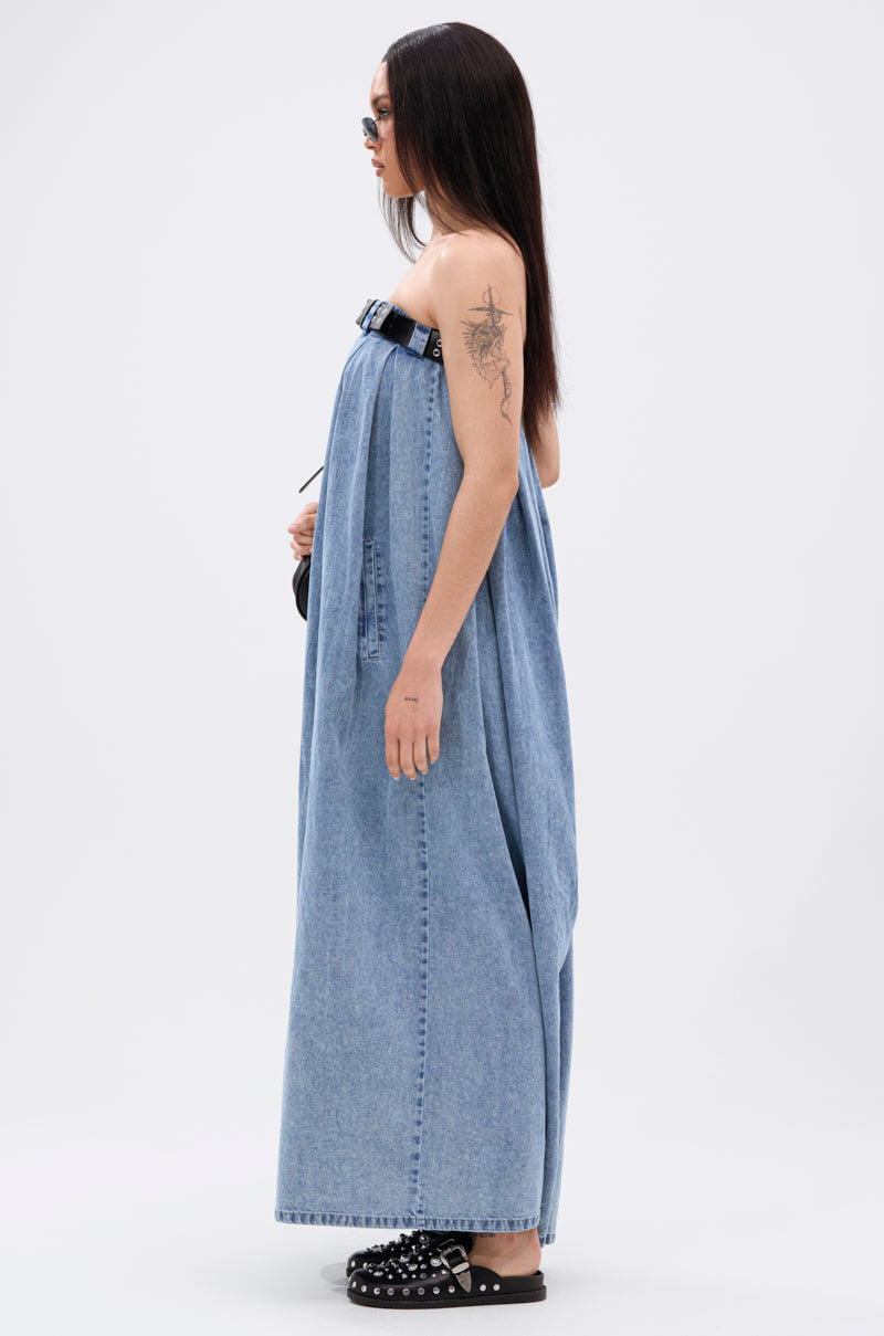 A woman with long dark hair and sunglasses models the DIANE STRAPLESS OVERSIZED JUMPSUIT IN DENIM, styled with a black buckle belt and studded sandals, standing in profile against a white background.