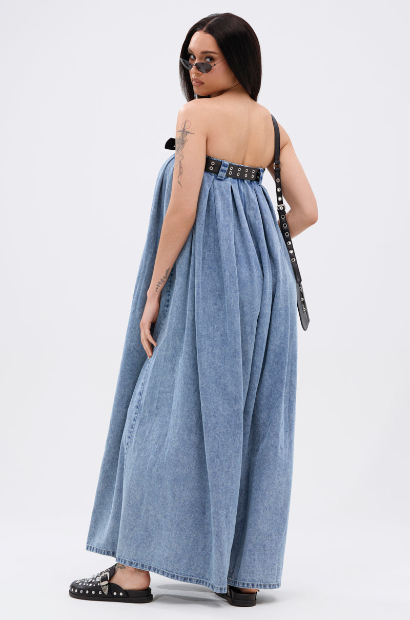 A woman in sunglasses models the DIANE STRAPLESS OVERSIZED JUMPSUIT IN DENIM with a black studded strap and sandals, standing backwards and looking over her shoulder against a plain white background.