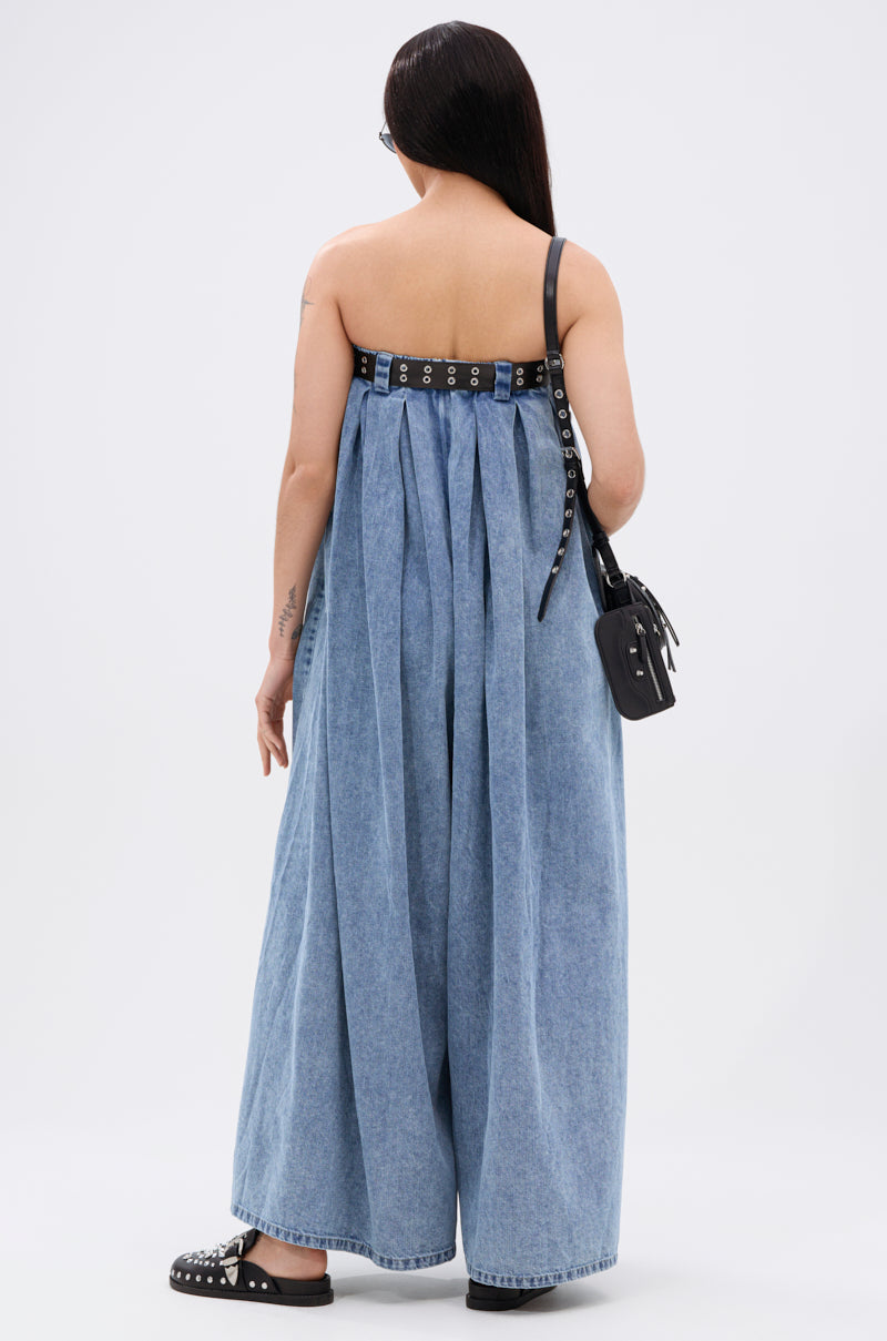 A person with long straight hair stands facing away in the DIANE STRAPLESS OVERSIZED JUMPSUIT IN DENIM, paired with black sandals, a black studded shoulder bag, and glasses, against a plain white background.