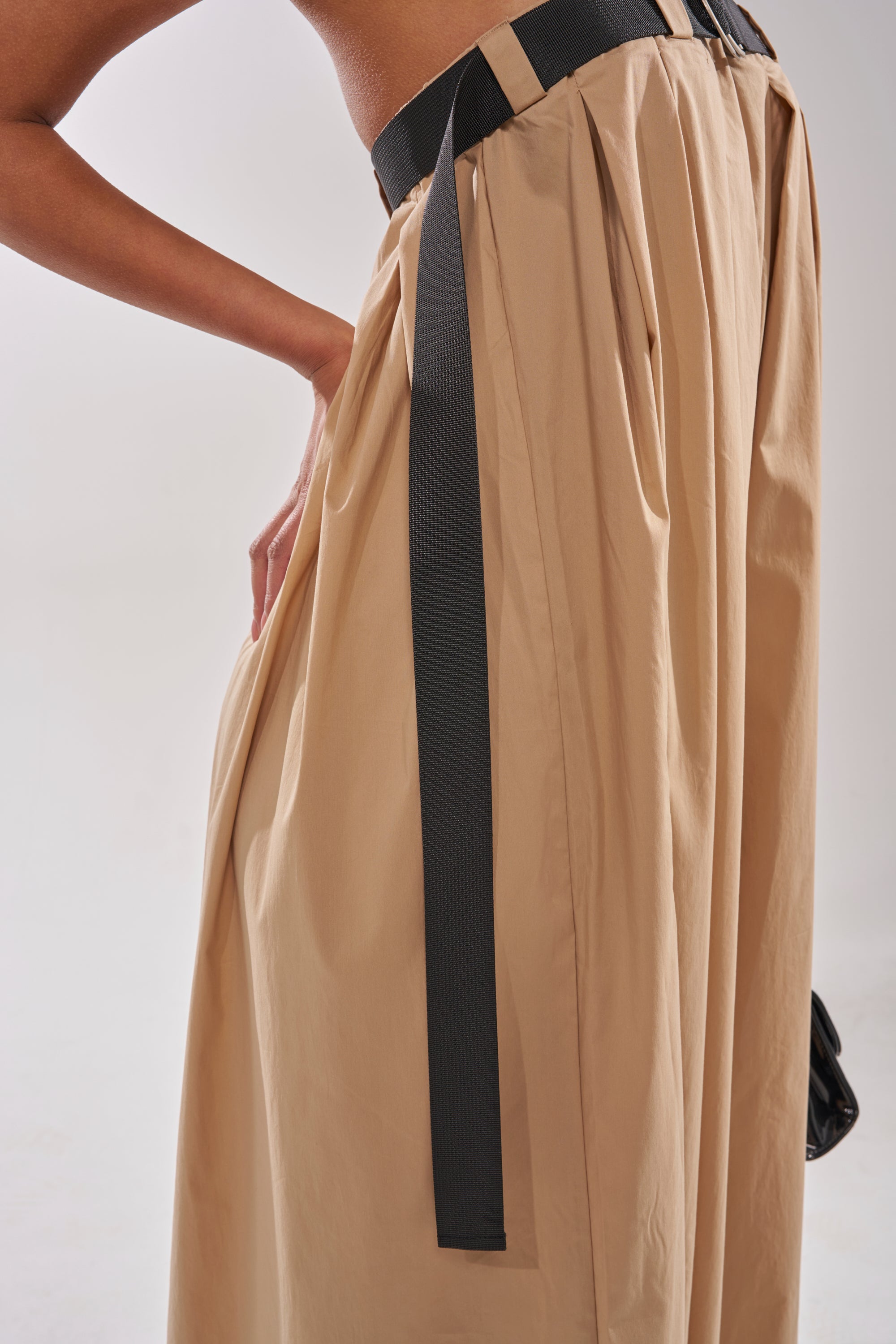A person wearing the DIANE STRAPLESS OVERSIZED KHAKI JUMPSUIT, featuring a high-waisted, pleated design with a wide black ribbon belt, stands against a plain light background with one hand on their hip. 9