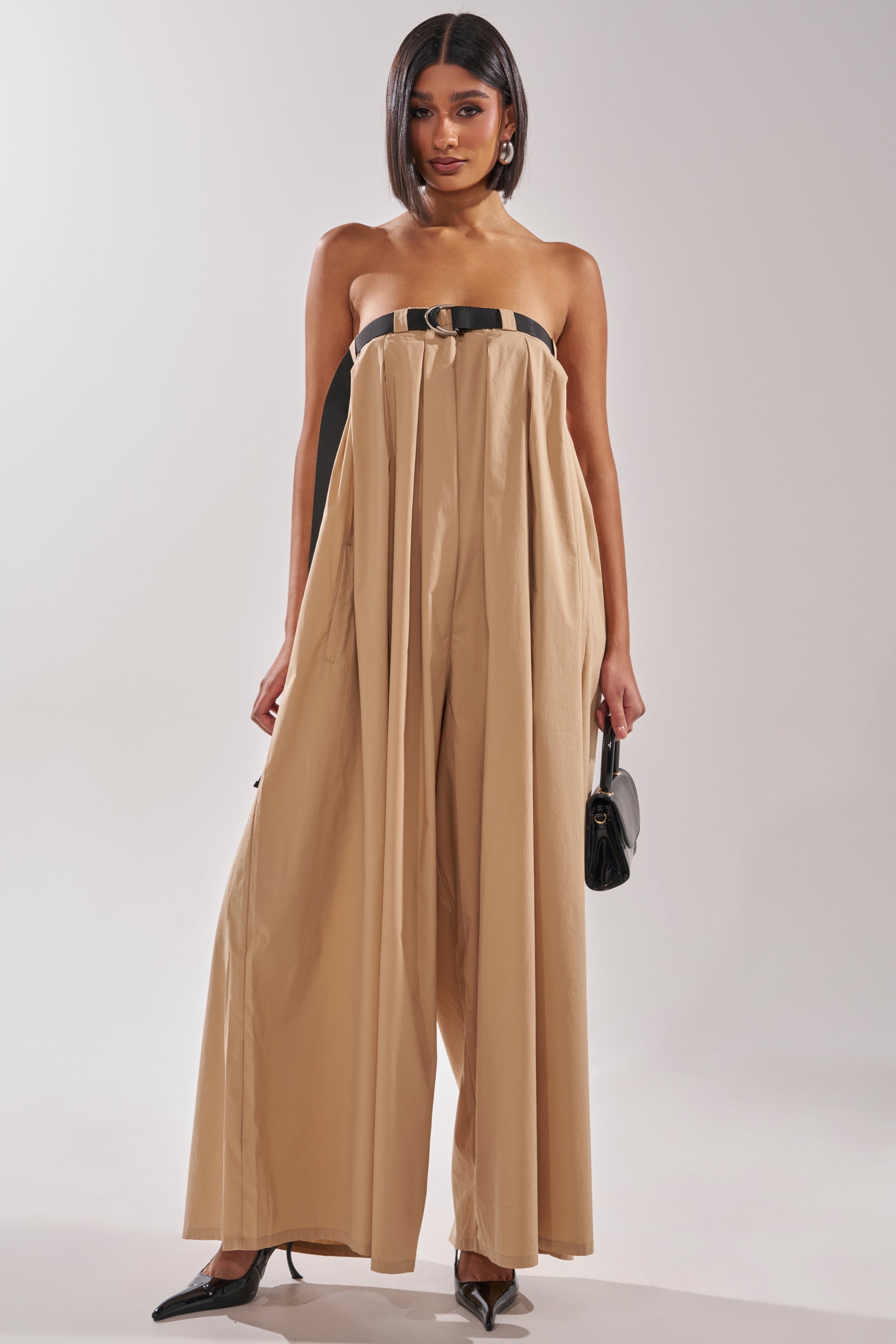 A woman confidently wears the DIANE STRAPLESS OVERSIZED KHAKI JUMPSUIT with a black belt, paired with a small black handbag and pointed black heels, set against a plain light background. 0