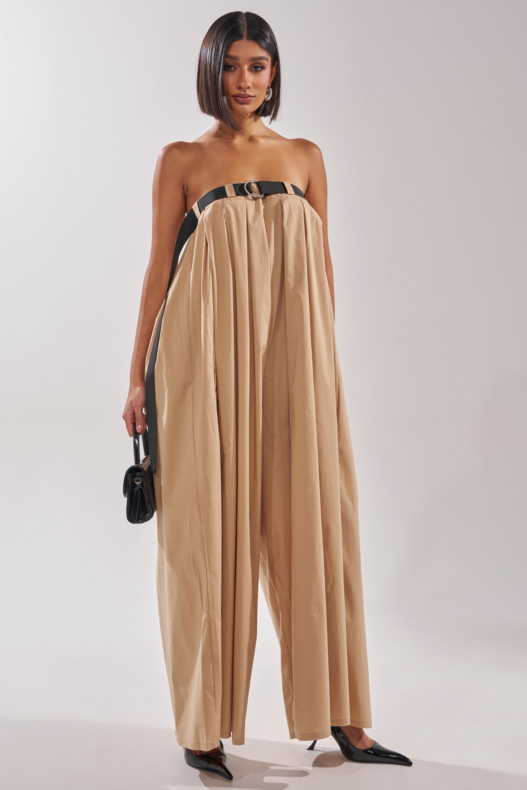 A woman wears the DIANE STRAPLESS OVERSIZED KHAKI JUMPSUIT with a black belt detail, styled with a small black purse and black pointed heels. The wide leg jumpsuit stands out against a plain, light-colored background. 1
