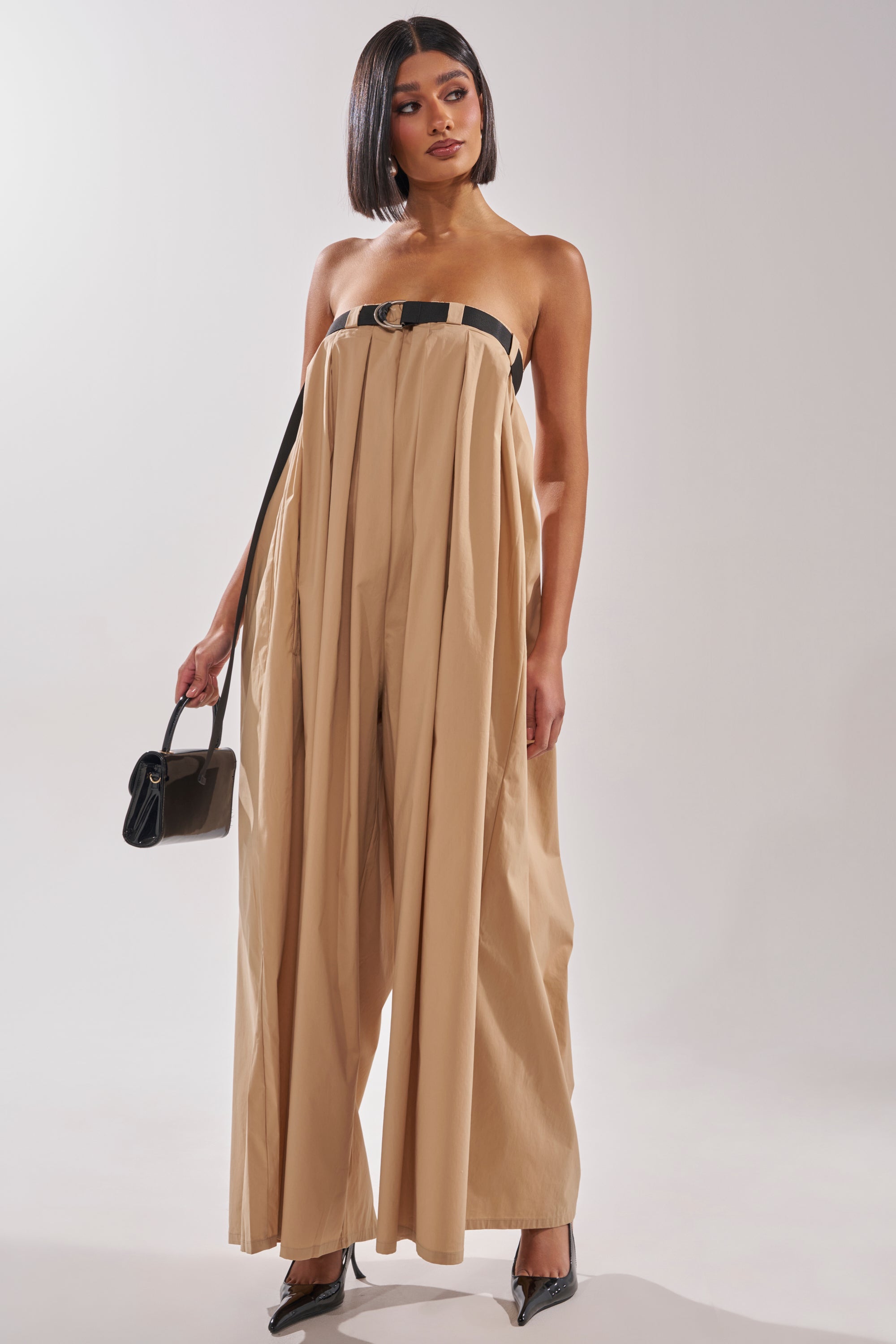 A woman models the DIANE Strapless Oversized Khaki Jumpsuit, featuring a black chest strap, paired with a small black handbag and pointed black heels, standing against a plain light background. 2
