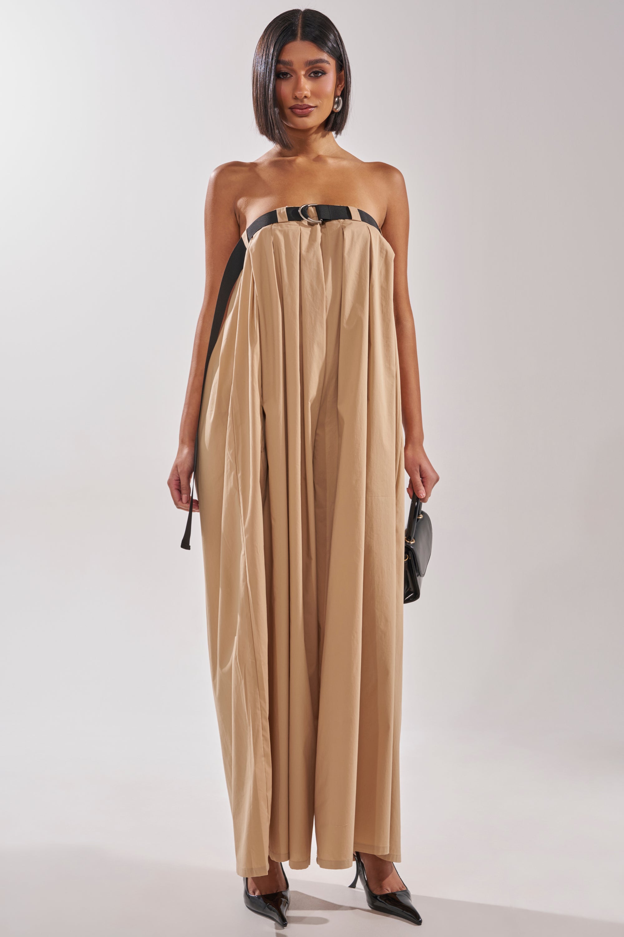 A woman wears the DIANE STRAPLESS OVERSIZED KHAKI JUMPSUIT with a black belt, holding a small black handbag and sporting pointed black heels. She has straight shoulder-length hair and a neutral expression. 3