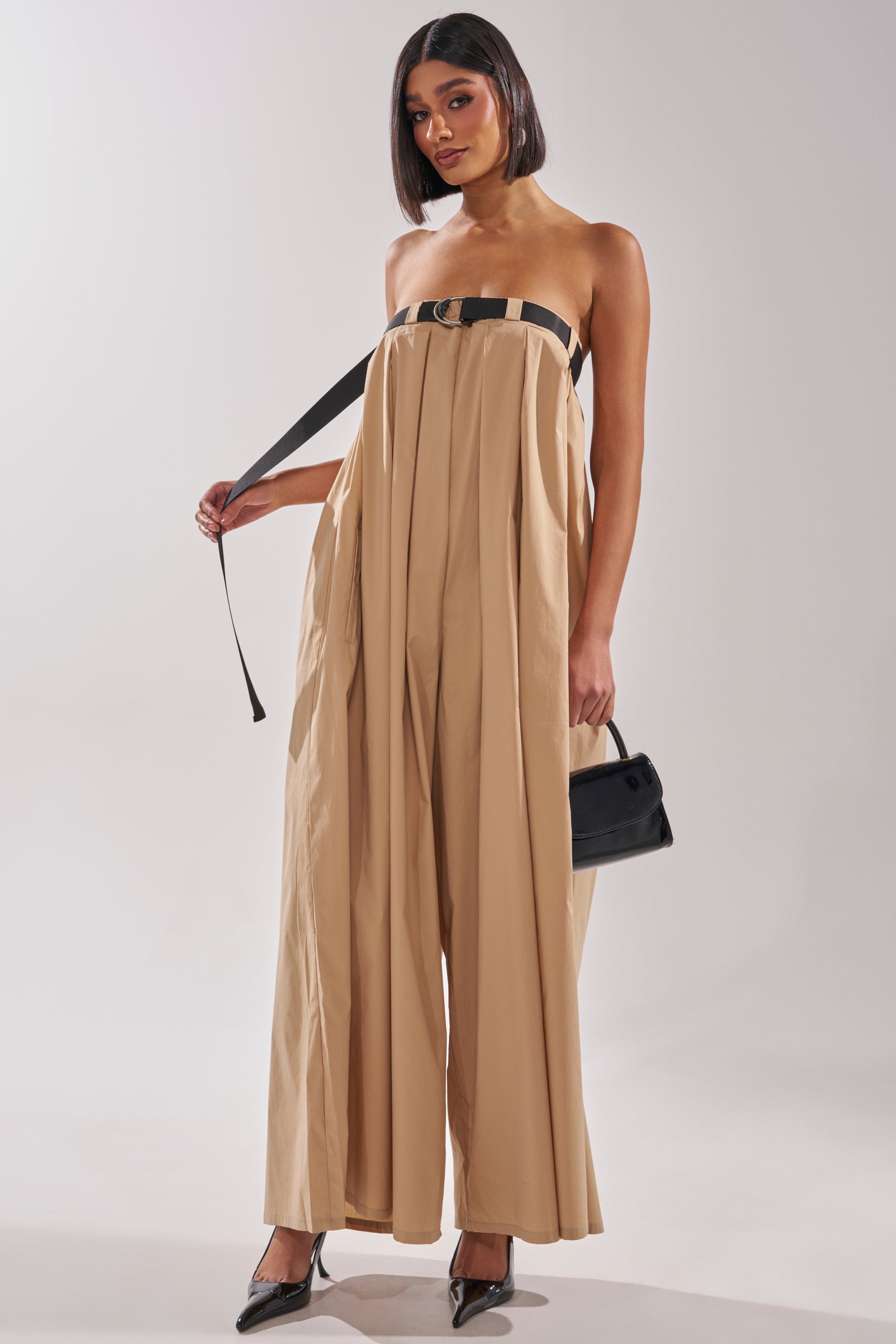 A woman wearing the DIANE STRAPLESS OVERSIZED KHAKI JUMPSUIT poses with a black handbag and black heels against a plain light background. 4