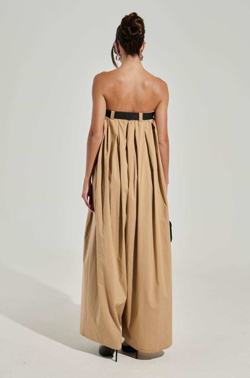 A person with a bun hairstyle is seen from the back, wearing the DIANE STRAPLESS OVERSIZED JUMPSUIT. The outfit is full-length and beige with a black belt, featuring a loose, flowing design akin to an oversized jumpsuit. The background is plain white-grey, and the individual stands on a flat surface.