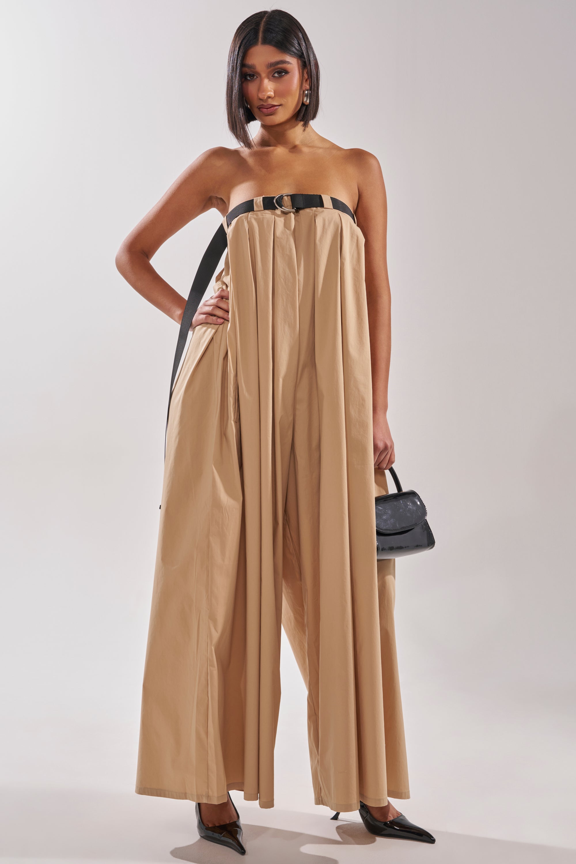 A woman poses against a plain light background in the DIANE STRAPLESS OVERSIZED KHAKI JUMPSUIT with wide legs, cinched at the waist with a black belt. She accessorizes with a small black handbag and sleek black pointed heels. 5