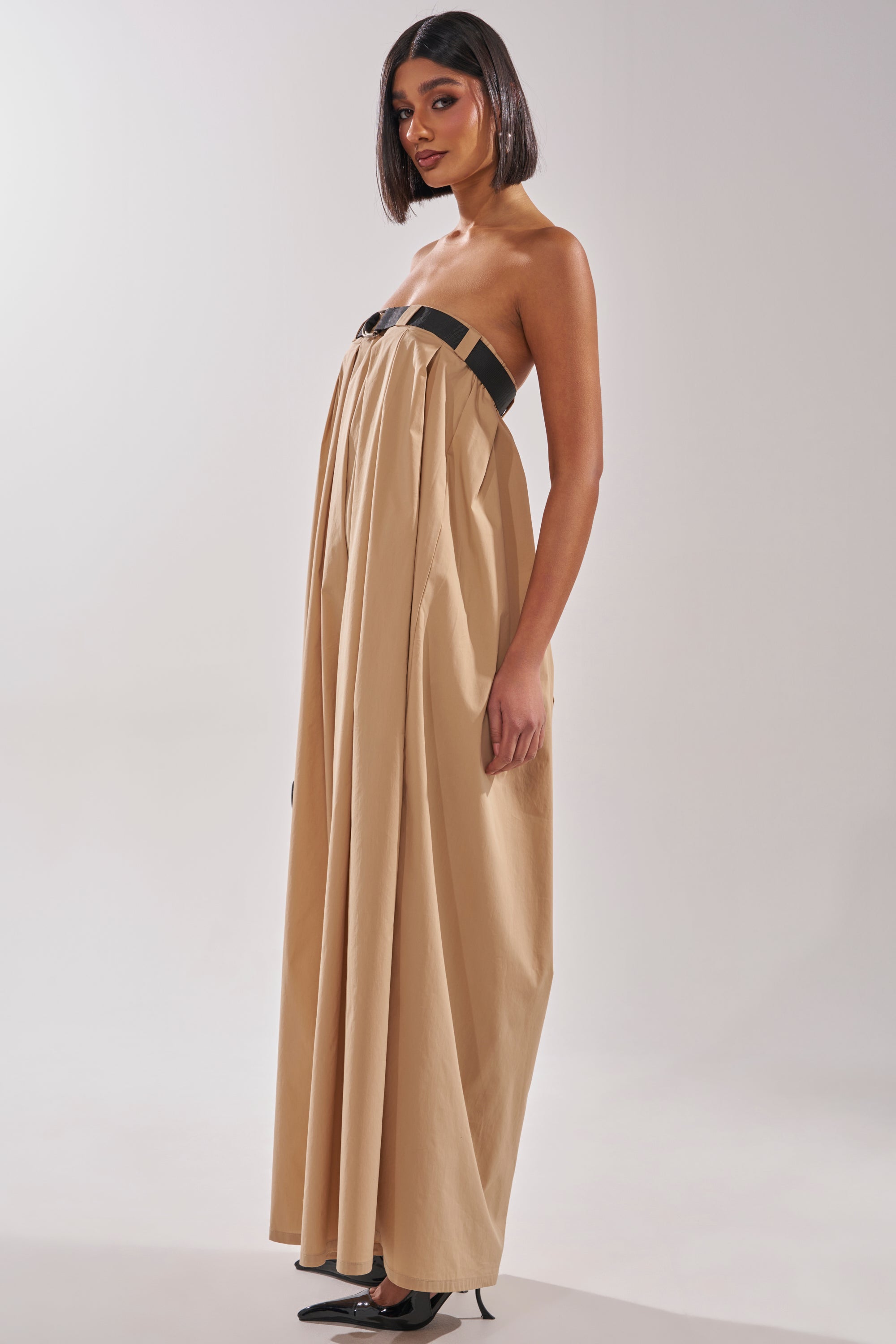 A woman wears the DIANE STRAPLESS OVERSIZED KHAKI JUMPSUIT with black accents, paired with black pointed-toe heels. She has straight, shoulder-length hair and stands against a plain light background. 6