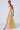 A woman wearing the MIMI COLORBLOCKED SCOOP NECK JUMPSUIT by JCR Trading LTD, which features vertical panels of green, pink, orange, and blue on the sides. She is standing against a plain white background, looking to her left. She also wears white high-heeled shoes and a gold necklace.