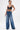 A person stands confidently in MY OWN THING STUDDED DENIM blue high-rise wide-leg jeans with chain details, a black leather crop top, and silver heels, holding a silver handbag. Tattoos and piercings are visible against a white background.