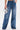 A person wears MY OWN THING STUDDED DENIM—high-rise, wide-leg blue jeans with silver studded details and large front pockets—paired with a shiny abstract silver handbag. The jeans graze the floor as sandals peek beneath.