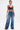 A woman with slicked-back hair wears the MY OWN THING STUDDED DENIM wide-leg jeans with silver chain details, paired with a black leather crop top, large hoop earrings, visible tattoos, a silver arm cuff, and a shiny silver purse.