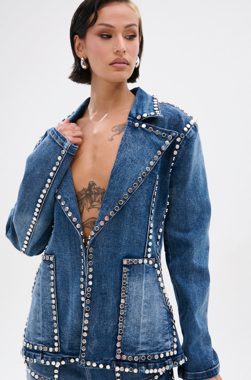 A person with short dark hair wears the MY OWN THING STUDDED DENIM BLAZER, featuring a notched lapel collar and tailored fit, unbuttoned to reveal a tattooed chest as they gaze to the side against a plain light background.