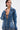 A person with short dark hair wears the MY OWN THING STUDDED DENIM BLAZER, featuring a notched lapel collar and tailored fit, unbuttoned to reveal a tattooed chest as they gaze to the side against a plain light background.