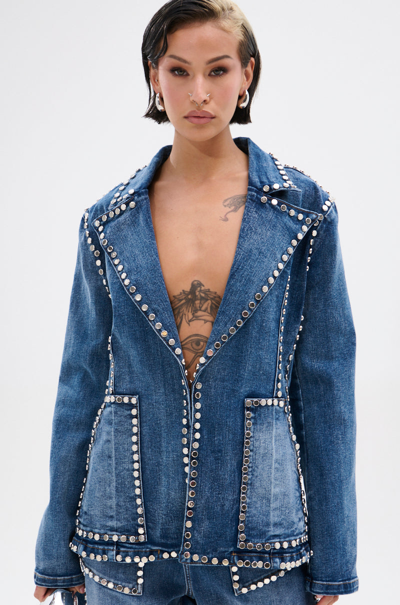 A person with short dark hair wears the MY OWN THING STUDDED DENIM BLAZER, featuring a tailored fit, deep V-neck, and notched lapel collar that reveals chest tattoos. The background is plain and light-colored.