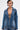 A person with short dark hair wears the MY OWN THING STUDDED DENIM BLAZER, featuring a tailored fit, deep V-neck, and notched lapel collar that reveals chest tattoos. The background is plain and light-colored.