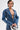 A person with short dark hair wears the MY OWN THING STUDDED DENIM BLAZER—tailored fit with a notched lapel and no shirt underneath, revealing a chest tattoo. They hold a shiny silver mask against a light plain background.