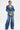 A person poses confidently in the MY OWN THING STUDDED DENIM BLAZER with a notched lapel, paired with wide-leg jeans and a metallic clutch. They have short dark hair, statement earrings, and chest tattoos against a white background.