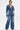 A woman with slicked-back hair poses against a white background in the MY OWN THING STUDDED DENIM BLAZER, paired with wide-leg jeans, studded high-heeled sandals, and a metallic handbag.