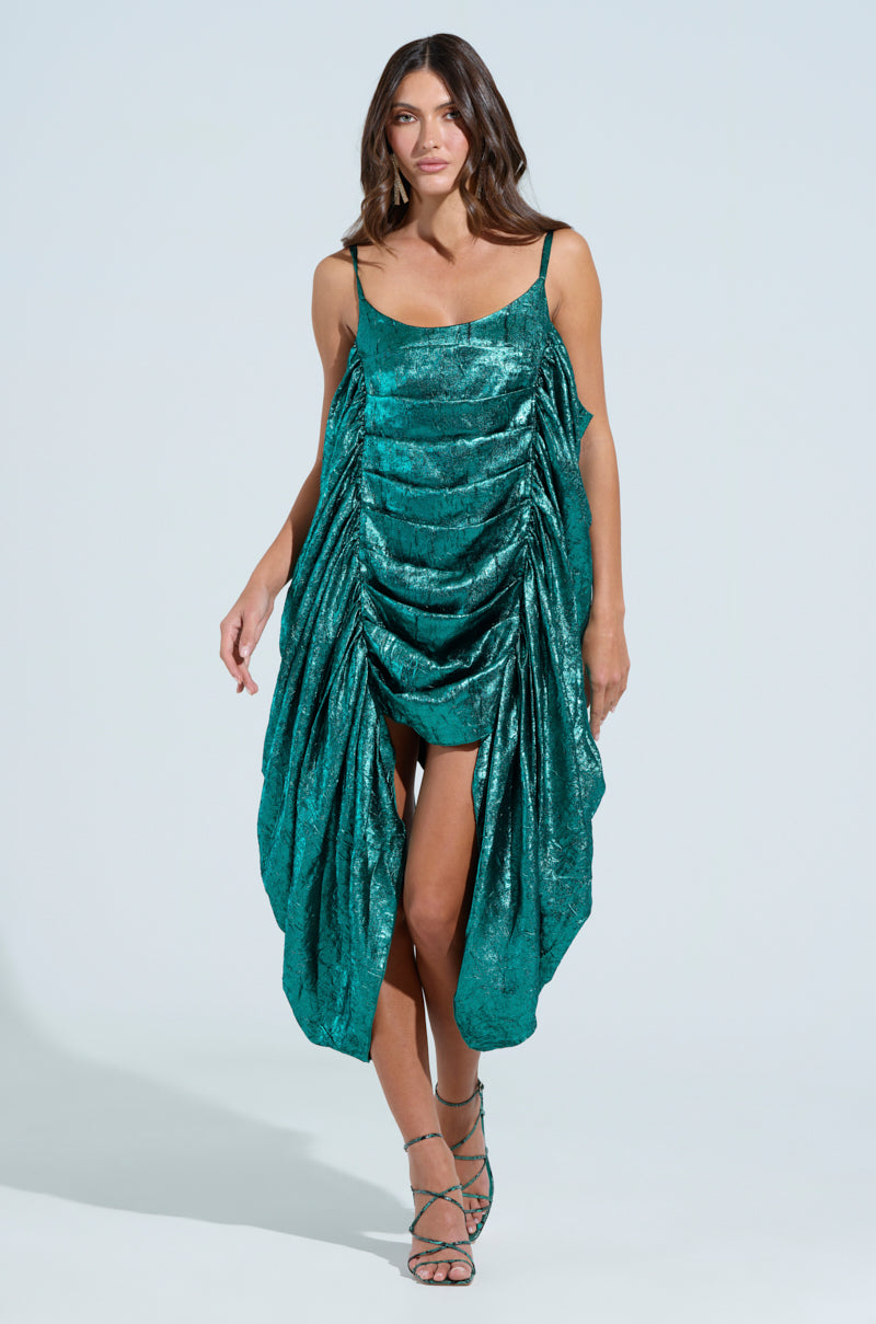 A woman models the Smooth Operator Ruched Midi Dress in metallic green, featuring a sleeveless design with ultra-ruched detailing and an asymmetrical hemline. She struts confidently in strappy sandals against a light background.