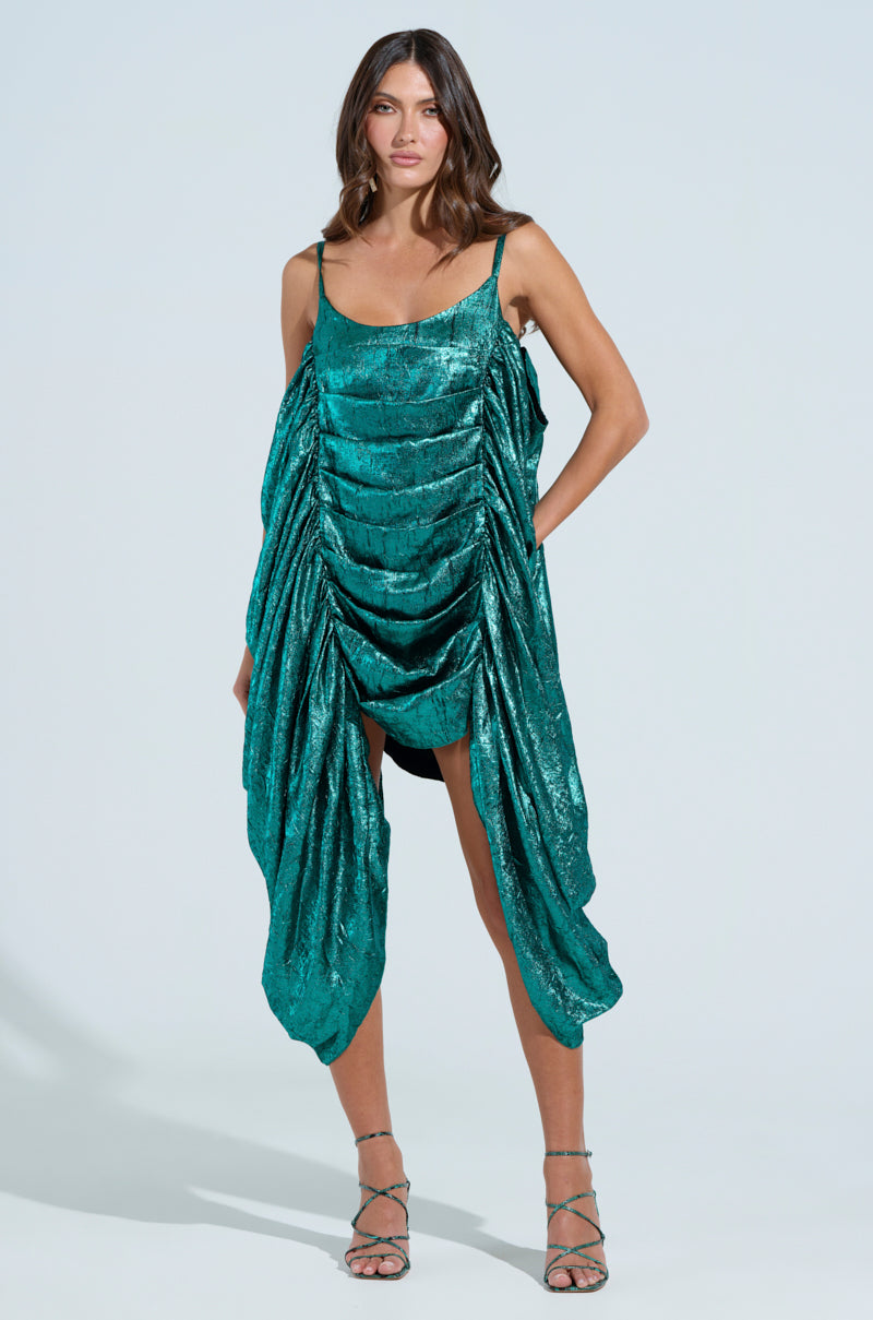 A woman elegantly poses in a shimmering teal Smooth Operator Ruched Midi Dress, featuring foiled metallic fabric, a draped ultra-ruched body, and thin straps. An asymmetrical hem complements her strappy high heels against the plain backdrop.