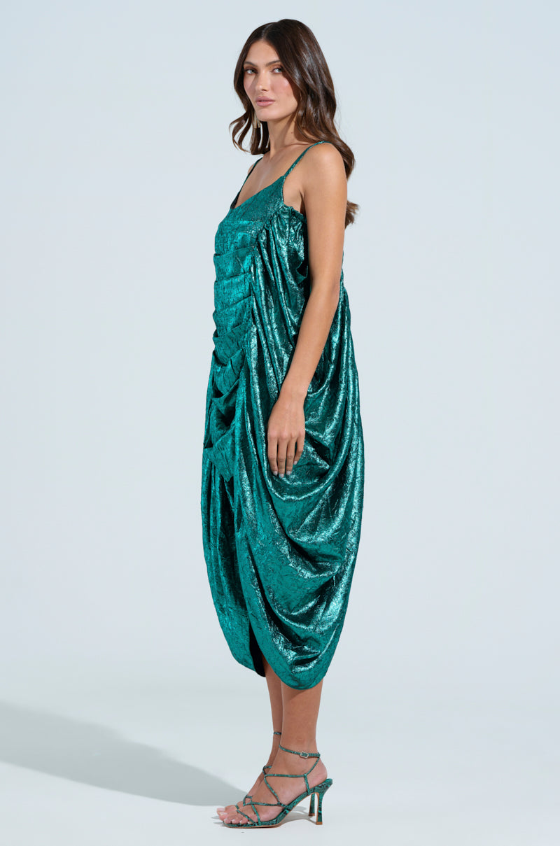 Wearing a shimmering emerald green Smooth Operator Ruched Midi Dress with spaghetti straps, a woman stands against a light background. The dress's ultra-ruched body detailing pairs perfectly with green strappy heels, and her long hair flows loosely.