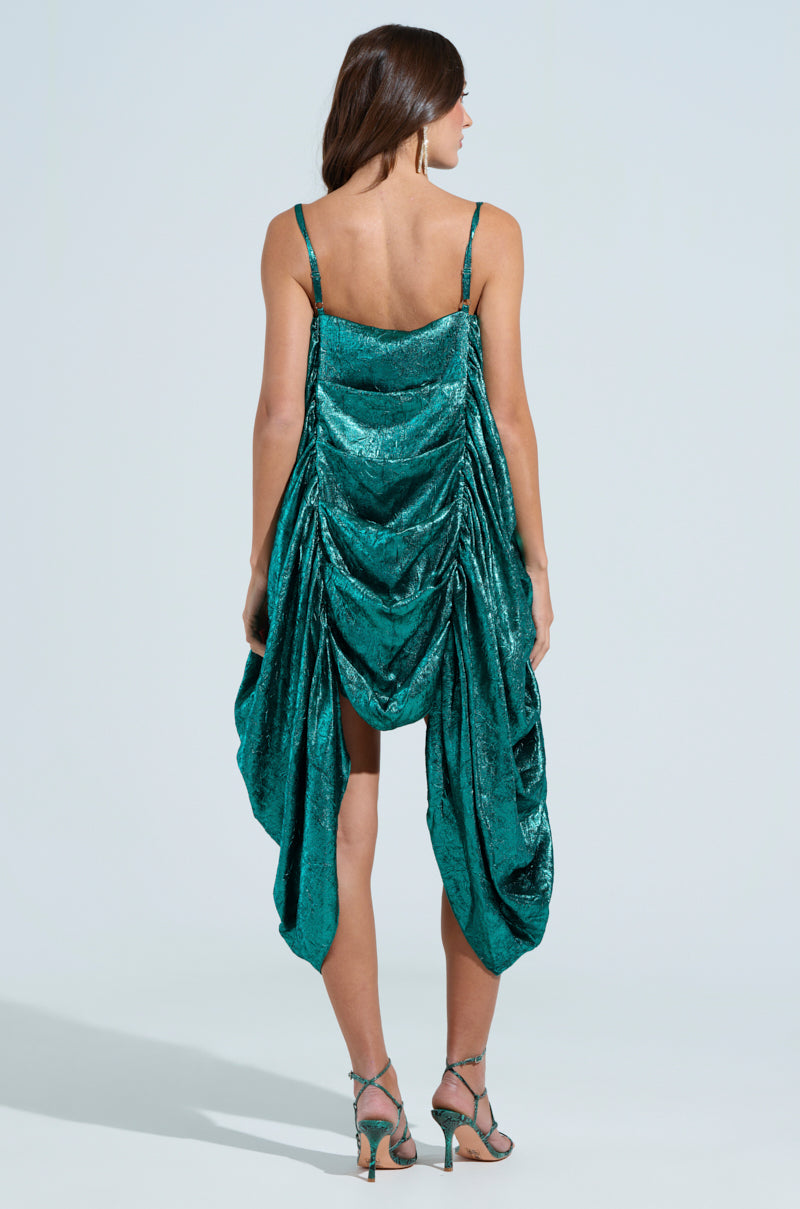 A woman with long brown hair elegantly wears a SMOOTH OPERATOR RUCHED MIDI DRESS in foiled metallic emerald green. The ultra-ruched body pairs with spaghetti straps as she poses on a light background, her high-heeled sandals adding the perfect touch.