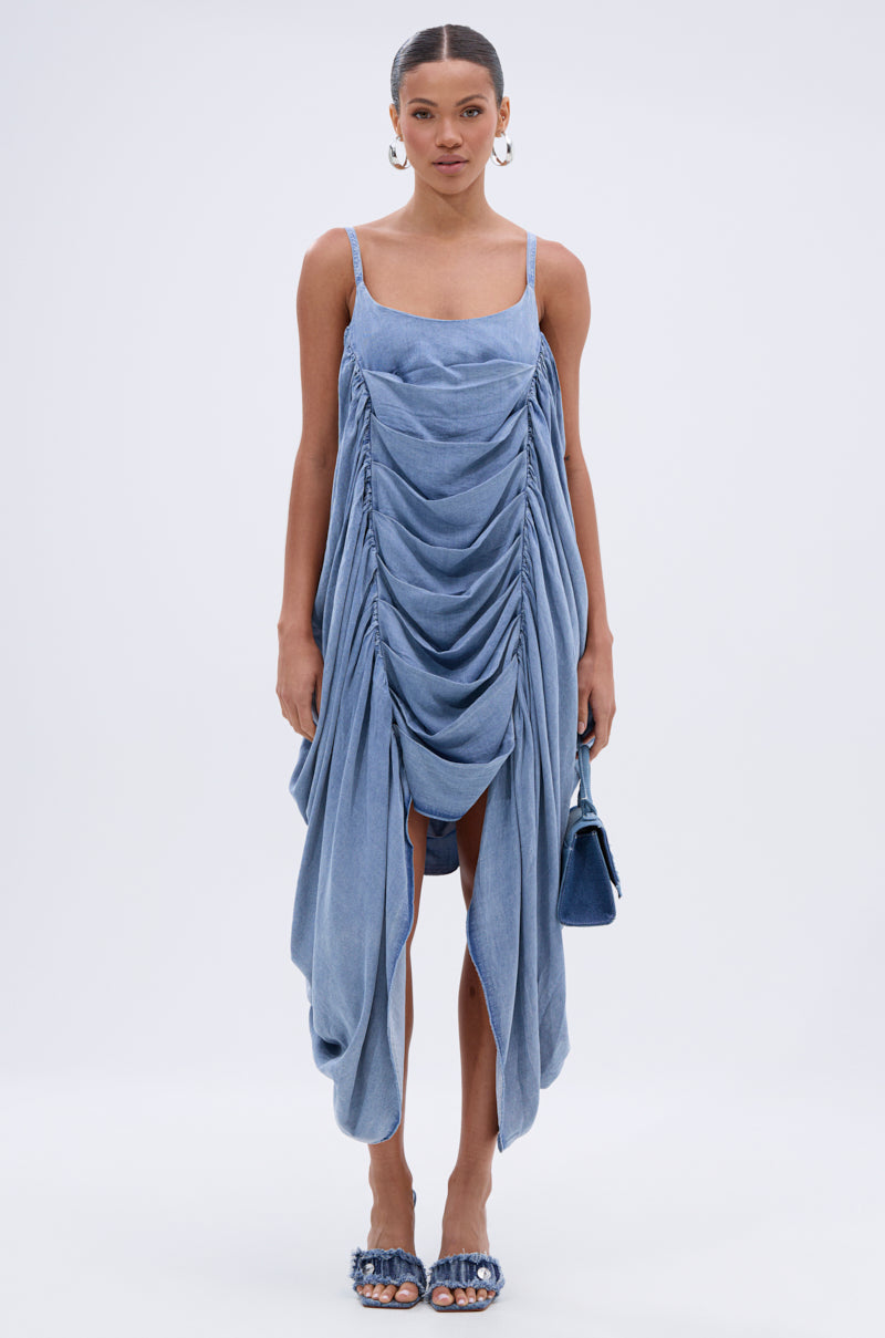 A woman faces forward in the SMOOTH OPERATOR RUCHED MIDI DRESS IN DENIM, featuring thin straps and a body-skimming fit. She pairs it with a matching purse, open-toe heels, gold hoop earrings, and wears her hair tied back against a white background.