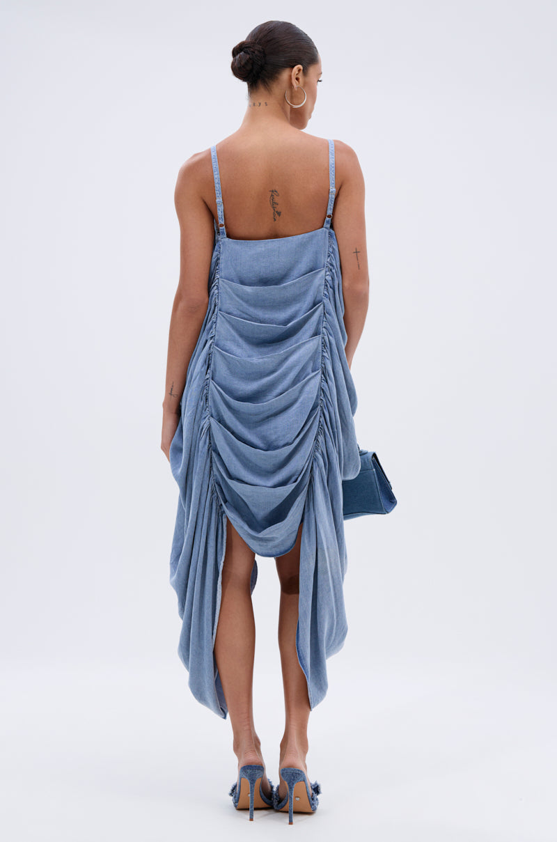 A woman, her hair in a neat bun and tattoos on her arms and back, wears the SMOOTH OPERATOR RUCHED MIDI DRESS IN DENIM with spaghetti straps, holding a matching blue bag and wearing blue heels.