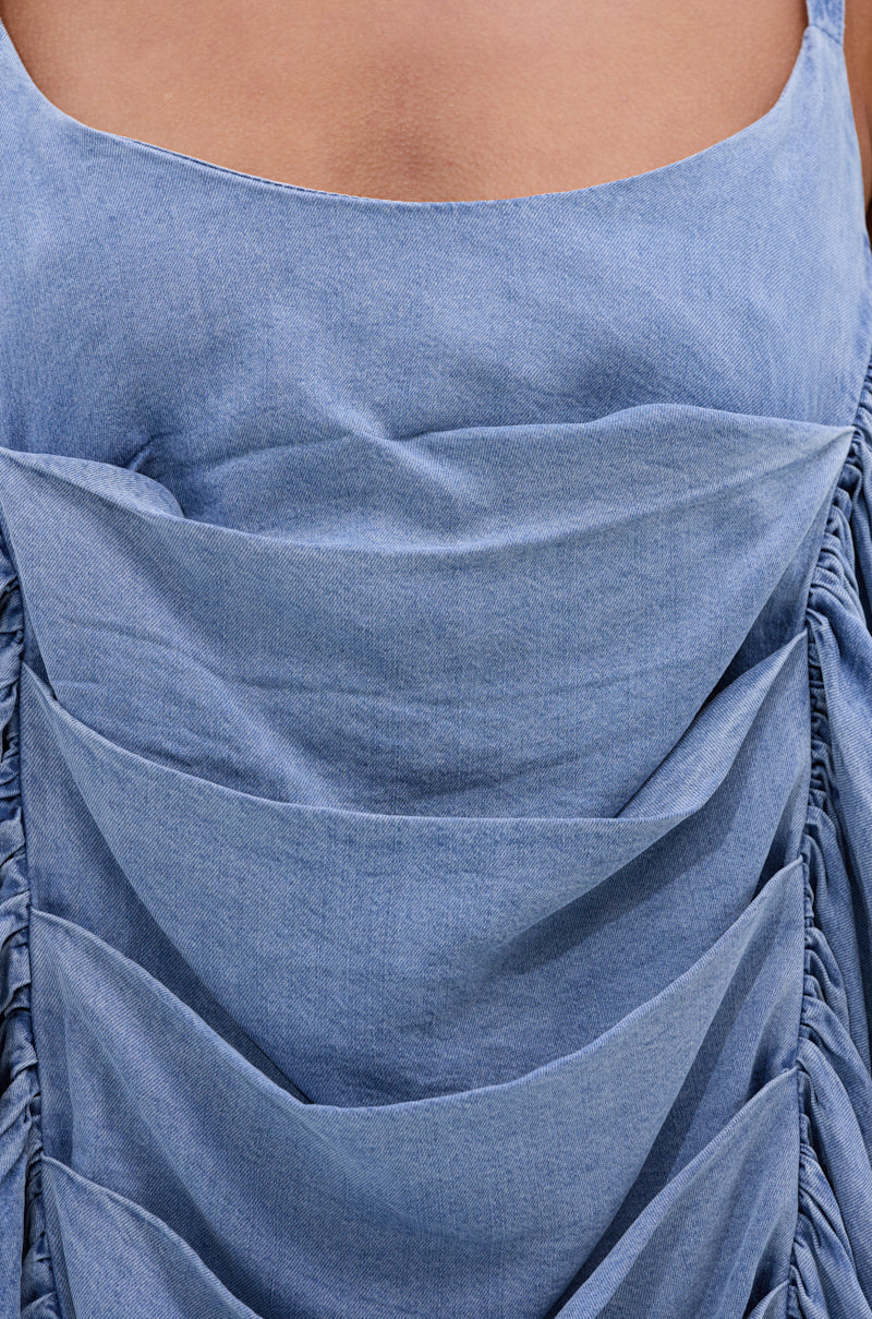 Close-up of the SMOOTH OPERATOR RUCHED MIDI DRESS IN DENIM, a light blue denim dress with ruched body detailing, gathered sections, exposed stitching, and adjustable tank straps, shown on a model.