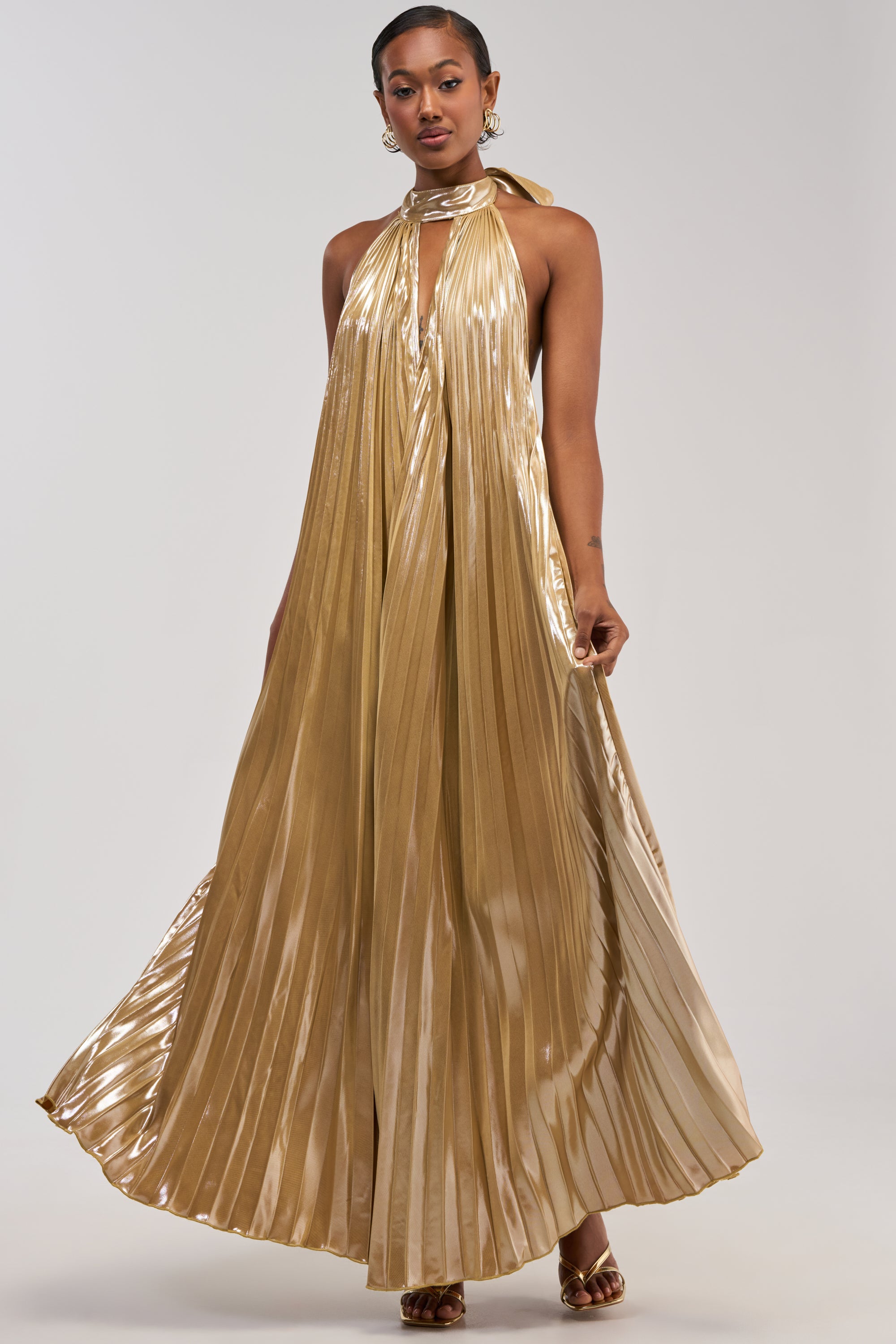 A woman stands gracefully in the DRIPPING IN GOLD SATIN MAXI DRESS, featuring a shiny gold hue, elegant pleats, and a halter-neck with a keyhole neckline as she slightly holds out the skirt. She completes her sophisticated look with gold earrings, a nose ring, and gold heels—making this ensemble a perfect choice for formal occasions or as an eye-catching wedding guest dress. The background is plain and light.