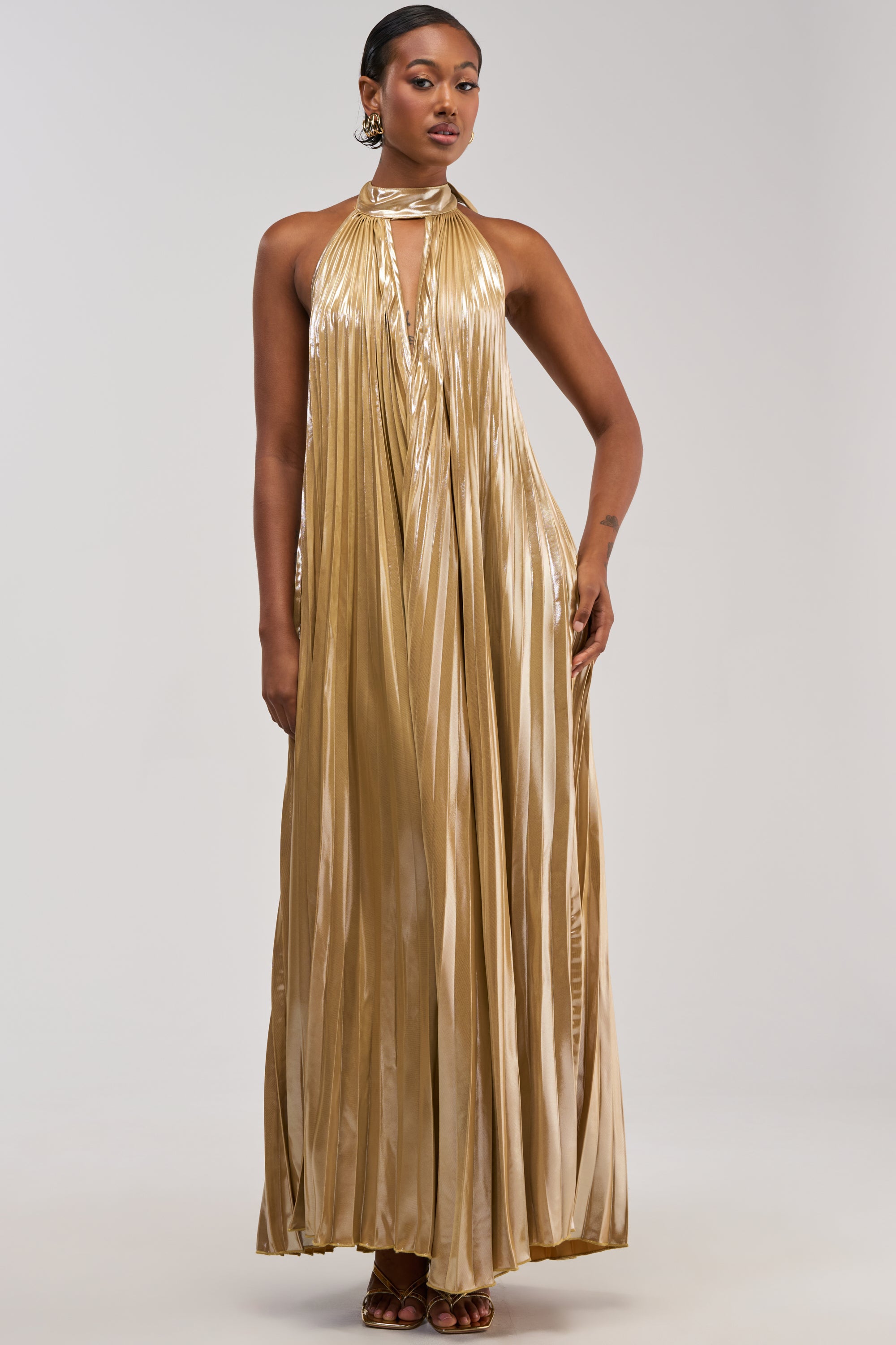 A woman stands in the DRIPPING IN GOLD SATIN MAXI DRESS, a long, metallic gold pleated maxi dress featuring a high neck and keyhole front. Styled as an elegant wedding guest look, she completes the outfit with gold earrings and brown heels against a plain light background, making it perfect for any formal occasion.