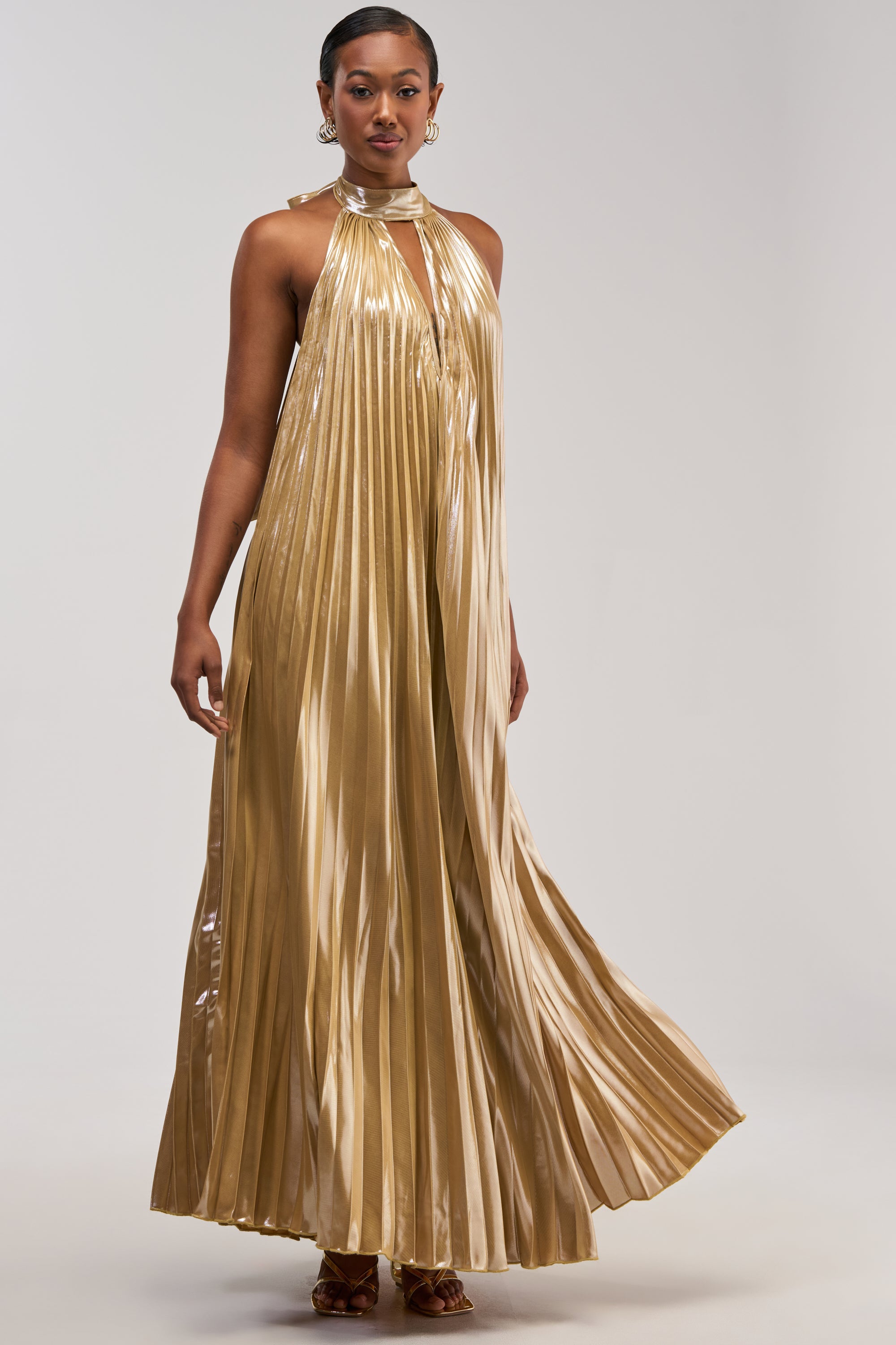 A woman stands confidently against a plain background, showcasing the DRIPPING IN GOLD SATIN MAXI DRESS—a stunning gold pleated halter-neck gown with a keyhole cutout, perfect for any formal occasion.