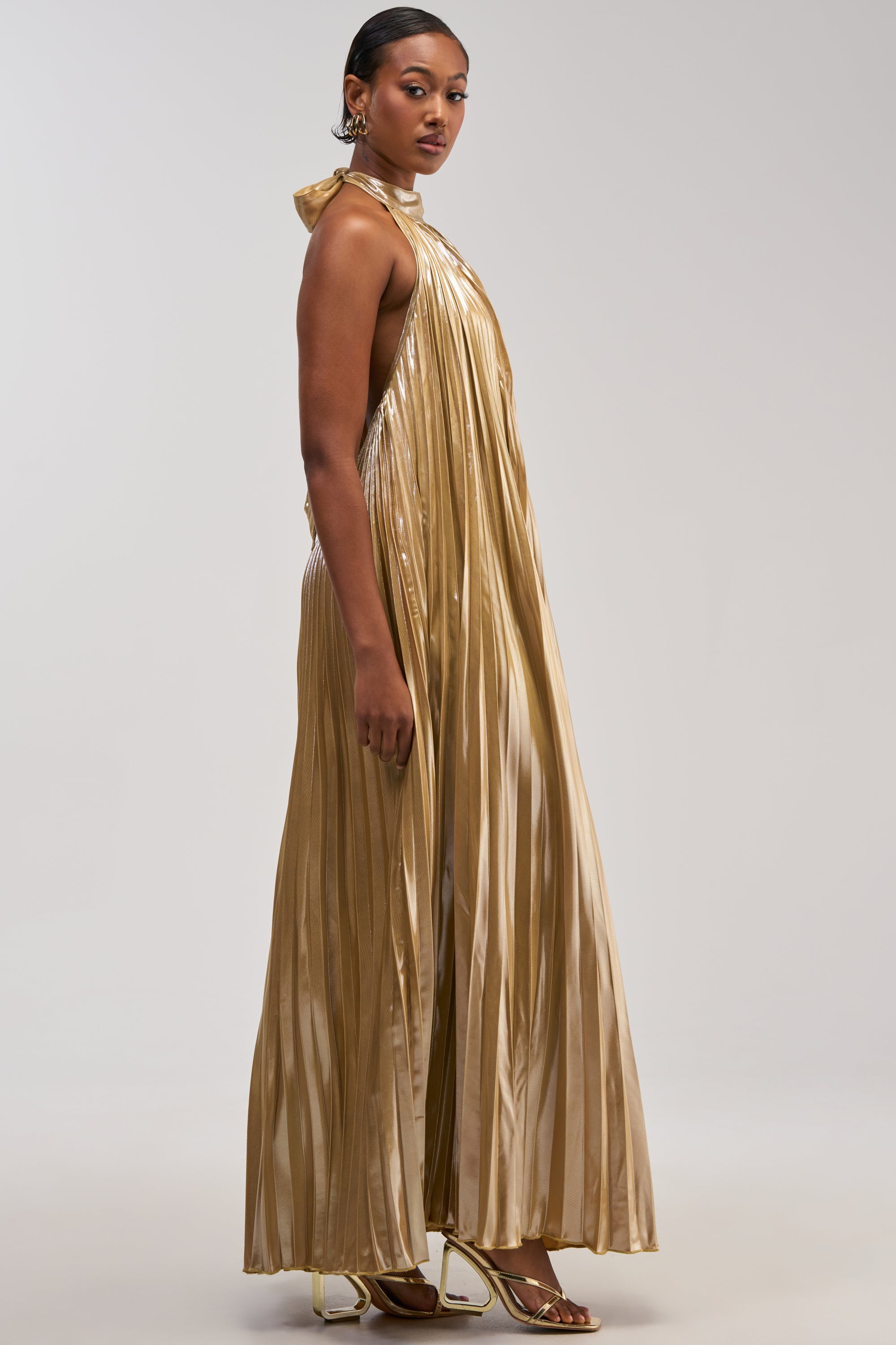 A woman stands in profile against a plain light background, wearing the DRIPPING IN GOLD SATIN MAXI DRESS—a shiny, gold pleated maxi dress featuring a halter neck and open back. She pairs this elegant wedding guest dress with gold heels and large earrings.