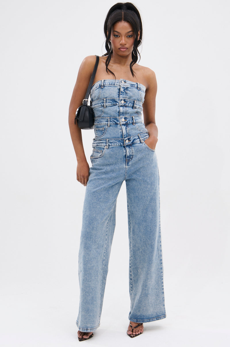 A woman poses against a plain background in the DENIM DENIM DENIM LAYERED JUMPSUIT, a strapless, belted denim design with wide legs. She pairs it with black heels, a small black shoulder bag, and wears her hair in loose waves.