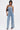 A woman poses against a plain background in the DENIM DENIM DENIM LAYERED JUMPSUIT, a strapless, belted denim design with wide legs. She pairs it with black heels, a small black shoulder bag, and wears her hair in loose waves.
