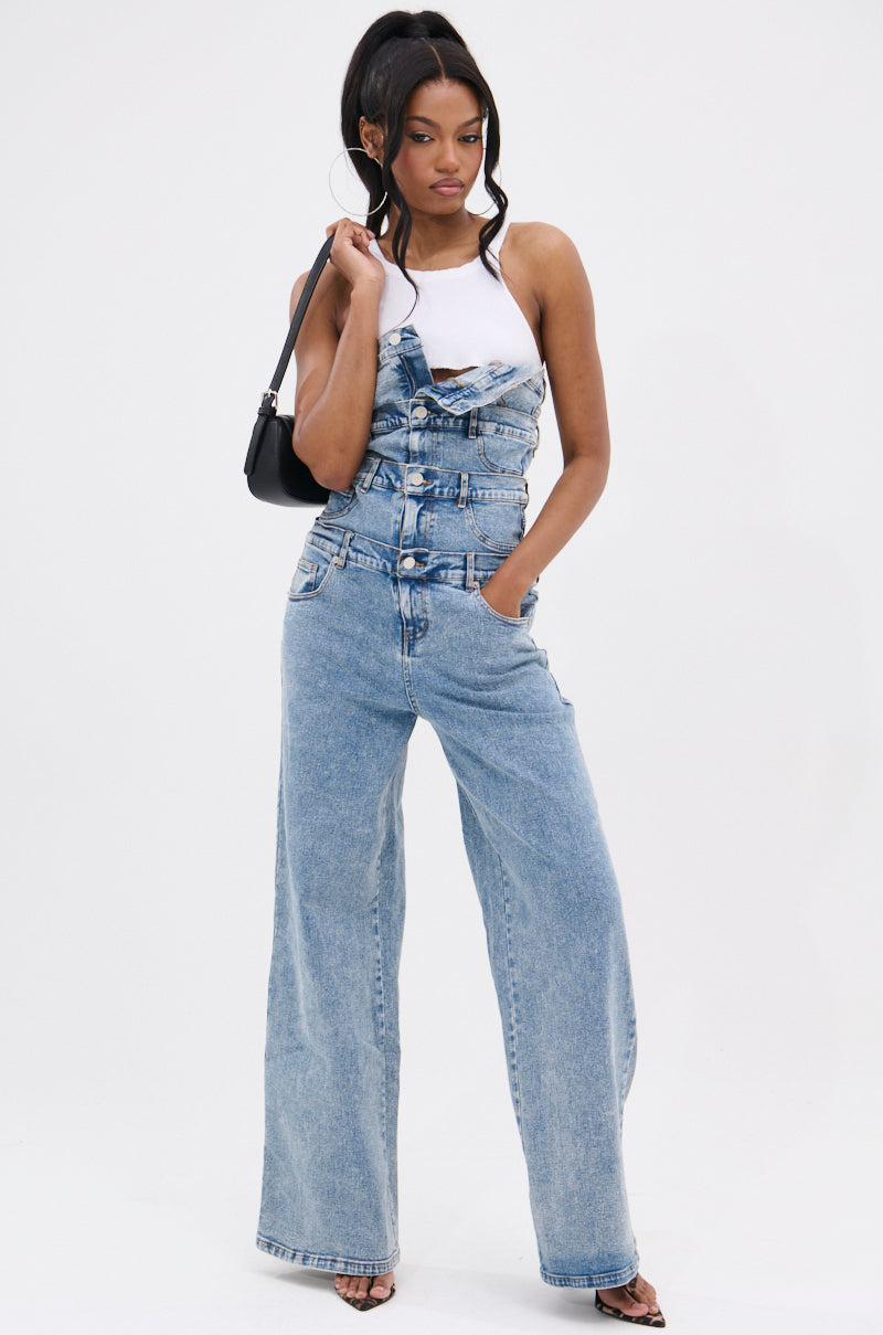 A woman stands confidently in the DENIM DENIM DENIM LAYERED JUMPSUIT with wide legs over a white sleeveless top. She has her hand in her pocket, carries a small black shoulder bag, and wears heeled sandals against a plain white background.