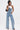 A woman stands confidently in the DENIM DENIM DENIM LAYERED JUMPSUIT with wide legs over a white sleeveless top. She has her hand in her pocket, carries a small black shoulder bag, and wears heeled sandals against a plain white background.