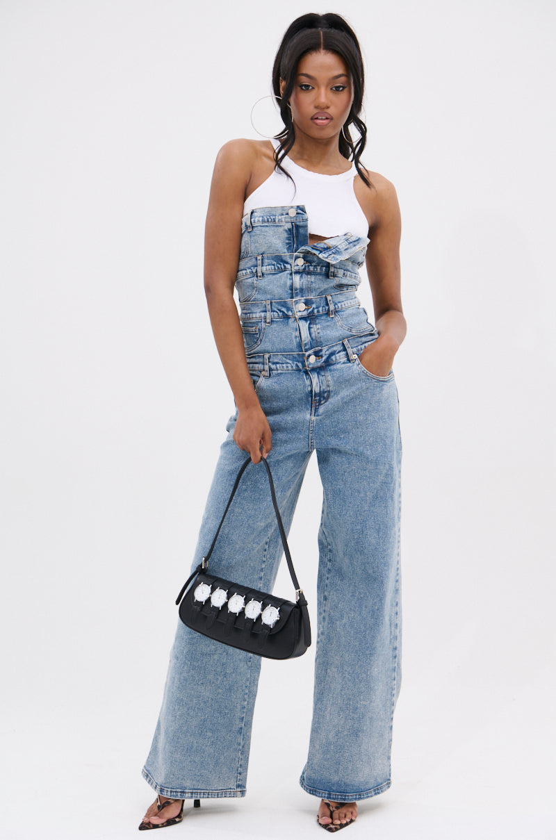 A woman stands confidently in the DENIM DENIM DENIM LAYERED JUMPSUIT, featuring a wide-leg silhouette and a strapless corset-style top over a white halter crop top. She holds a black handbag and wears black open-toe heels against a white background.