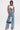 A woman stands confidently in the DENIM DENIM DENIM LAYERED JUMPSUIT, featuring a wide-leg silhouette and a strapless corset-style top over a white halter crop top. She holds a black handbag and wears black open-toe heels against a white background.